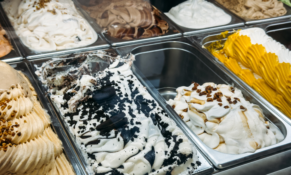 Uma vitrine de gelateria com vários sabores de sorvete expostos em cubas de inox. Há opções como chocolate, creme, cookies & cream e outros sabores variados, alguns decorados com pedaços de biscoito, caldas ou granulados, destacando a diversidade e o apelo visual.