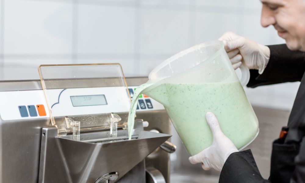 A imagem mostra a etapa de fabricação. Um profissional uniformizado e usando luvas despeja uma mistura líquida (aparentemente de pistache ou ervas, devido à cor verde clara) em uma máquina de sorvete industrial.