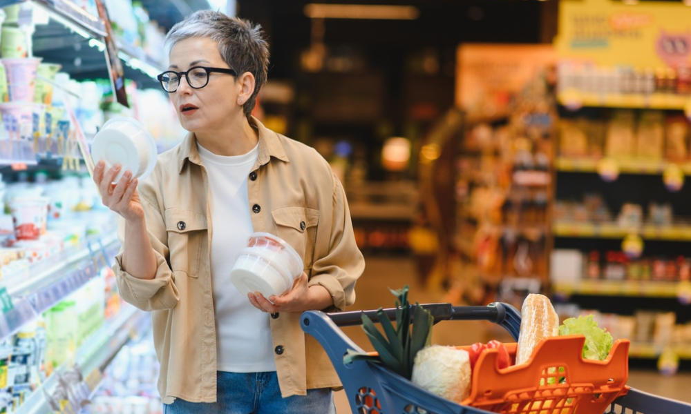 Uma mulher madura, com óculos e cabelos grisalhos, compara dois potes de produtos refrigerados (como laticínios ou pastas). Ela simboliza a preocupação com a qualidade e a escolha consciente em relação à saúde.