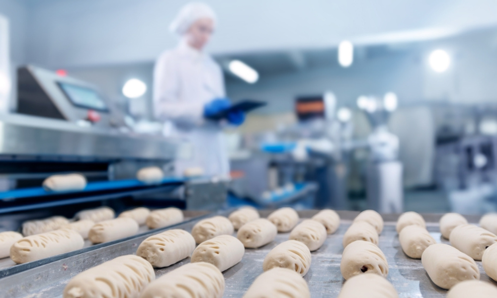 Várias fileiras de pães ou massas cruas em uma esteira metálica de uma fábrica. Ao fundo, uma funcionária vestindo uniforme branco, touca e luvas azuis opera um painel de controle ou tablet.