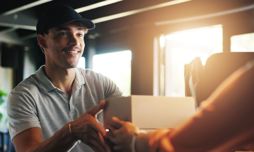 Um entregador sorridente, vestindo boné preto e camisa polo cinza, entregando uma caixa de papelão branca para uma pessoa (da qual vemos apenas o braço).