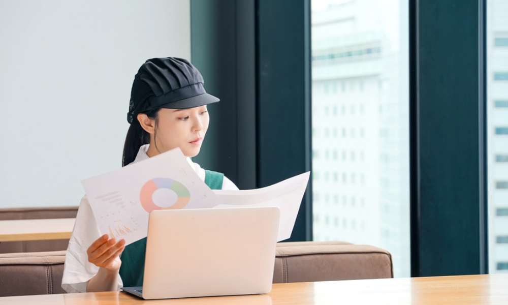 Uma mulher jovem, vestindo uniforme (avental verde e uma boina de estilo culinário), sentada à frente de um notebook em um ambiente moderno e bem iluminado.