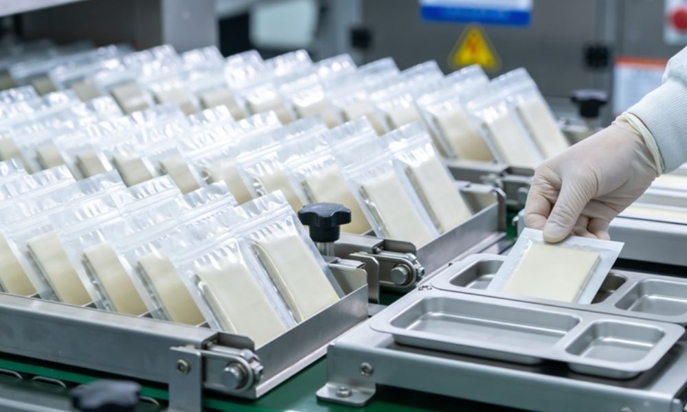 Linha de produção industrial onde várias embalagens plásticas transparentes com alimentos fatiados avançam organizadas em uma esteira metálica. À direita, uma mão com luva branca manipula uma das embalagens.