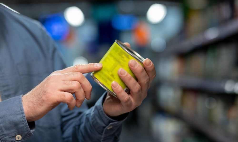 Close-up nas mãos de uma pessoa vestindo camisa azul que segura uma lata de alumínio com rótulo verde-limão. O dedo indicador aponta para as informações do rótulo, tendo o corredor desfocado de um supermercado ao fundo.