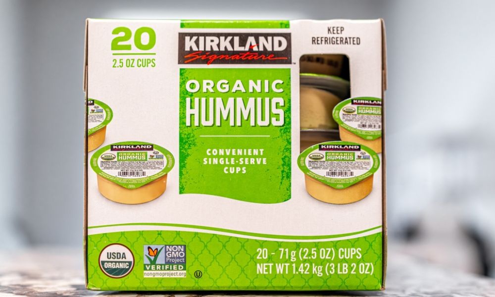 Uma embalagem de papelão branca e verde da marca Kirkland contendo 20 potes individuais de 71g de homus orgânico (Organic Hummus). A caixa exibe selos de produto orgânico e livre de transgênicos em fundo claro e desfocado.