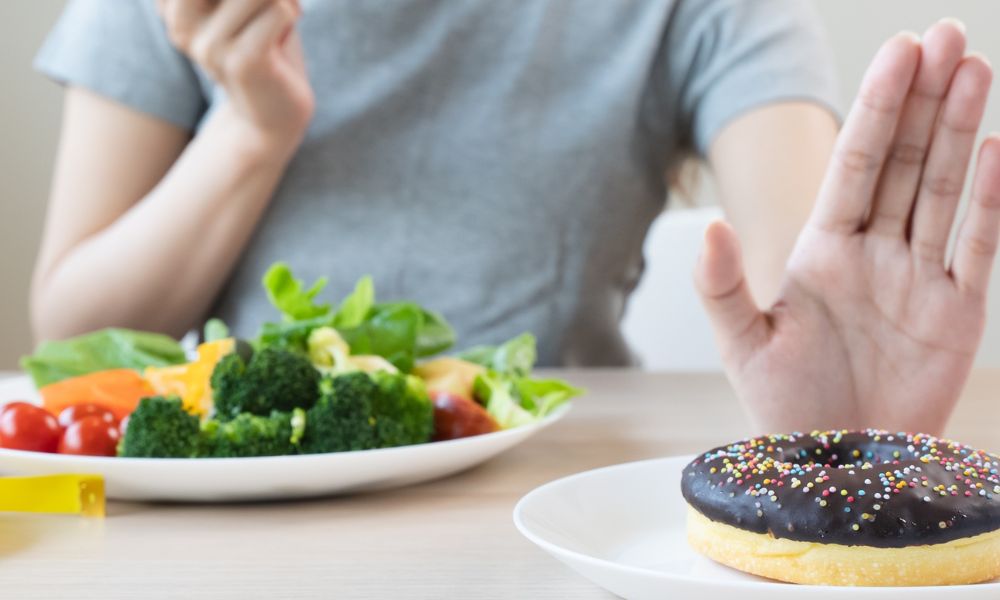 Uma mão estendida faz um gesto de recusa em direção a um donut com cobertura de chocolate e granulados em primeiro plano. Ao fundo, um prato com brócolis, tomates e vegetais saudáveis aparece ao lado de uma pessoa desfocada.