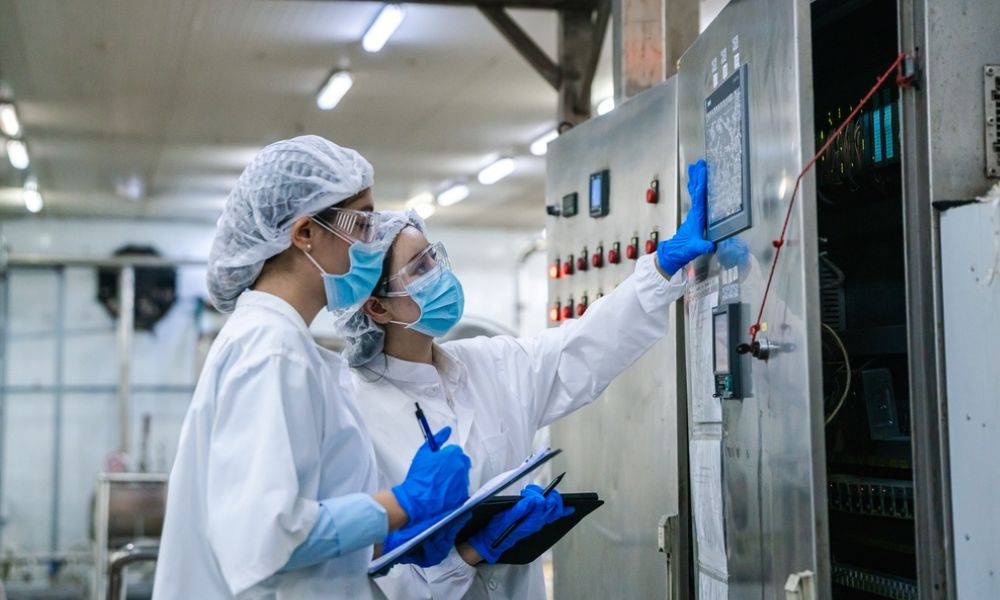 Duas profissionais de jaleco, máscara e touca operando e checando dados em um painel de controle industrial de uma fábrica de alimentos.