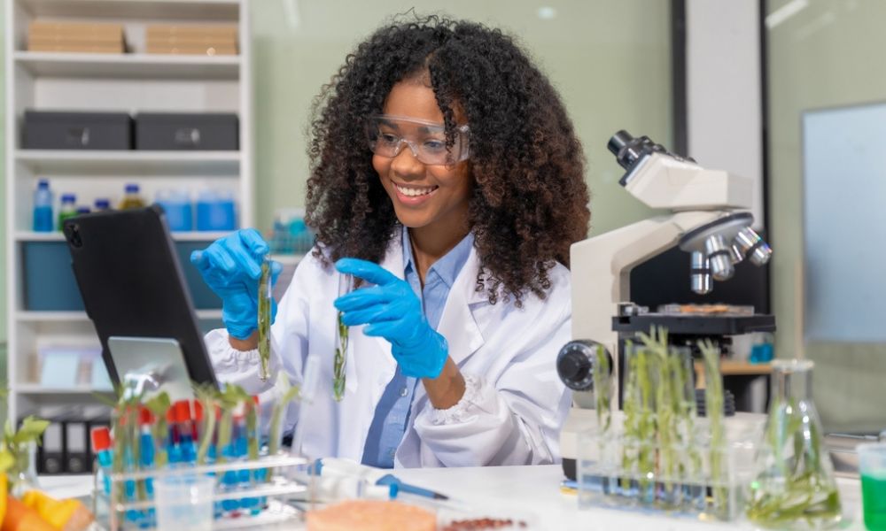 Uma cientista sorridente em um laboratório botânico, analisando amostras de plantas em tubos de ensaio com o auxílio de um microscópio e um tablet.