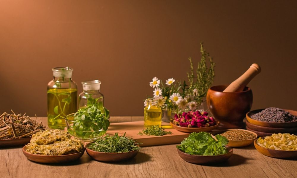 Uma bancada rústica de madeira organizada com frascos de óleos essenciais, ervas frescas, flores de camomila e um almofariz com pistilo para moagem.