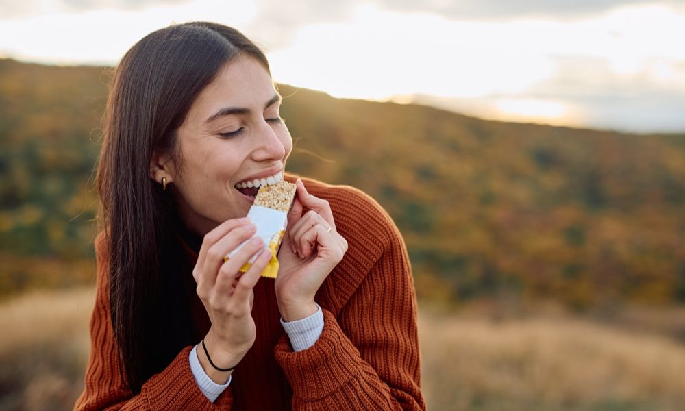 Uma mulher sorridente vestindo suéter marrom de tricô, comendo uma barra de cereal ou de proteína ao ar livre com paisagem outonal ao fundo.
