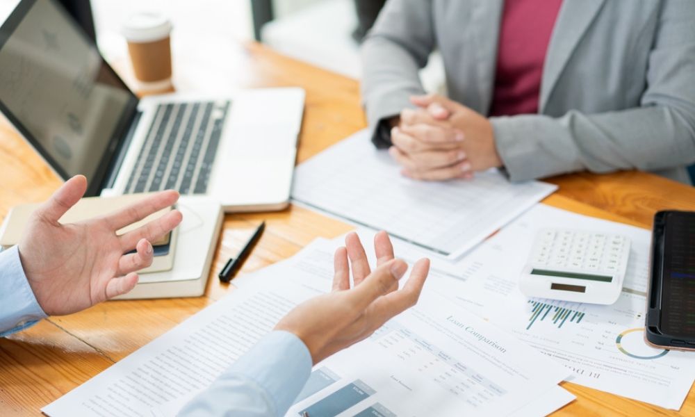 Pessoas reunidas ao redor de uma mesa de madeira com papéis, calculadora e notebook, conversando e gesticulando sobre documentos de comparação financeira.