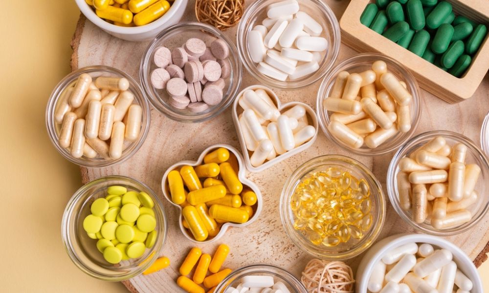 Diversas tigelas de vidro e potinhos dispostos sobre uma tábua de madeira com variados tipos de comprimidos, pílulas e cápsulas coloridas (amarelas, brancas, verdes e rosa).