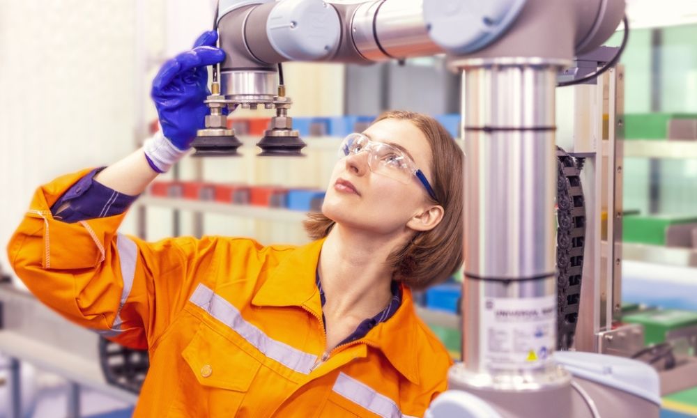 Uma técnica/engenheira usando EPI (óculos de proteção, luvas azuis e macacão de trabalho laranja) inspecionando a ferramenta de ponta de um robô colaborativo.