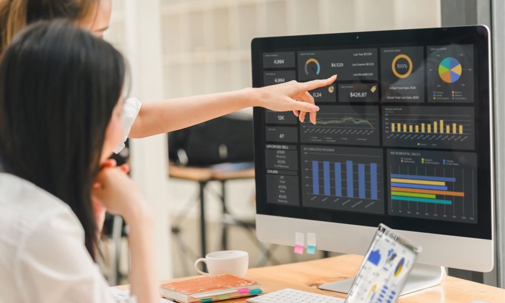 Mulher apontando para a tela de um computador que exibe gráficos, métricas e dashboards de desempenho financeiro e de vendas.