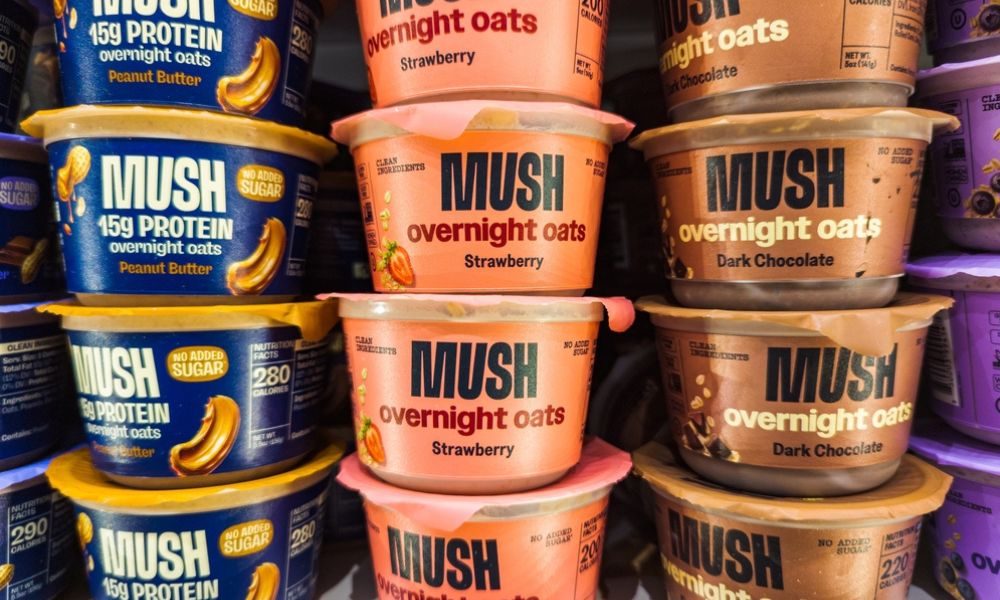 Prateleira de supermercado empilhada com recipientes do produto MUSH Overnight Oats nos sabores Peanut Butter (azul), Strawberry (laranja) e Dark Chocolate (marrom).