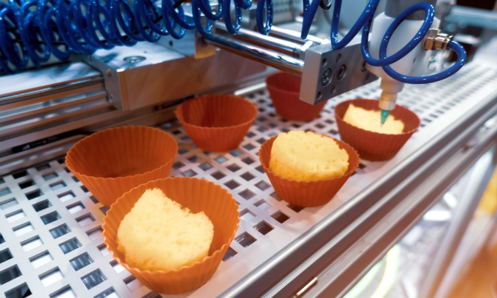 Maquinário automatizado recheando ou decorando pequenos bolos em forminhas alaranjadas numa esteira.