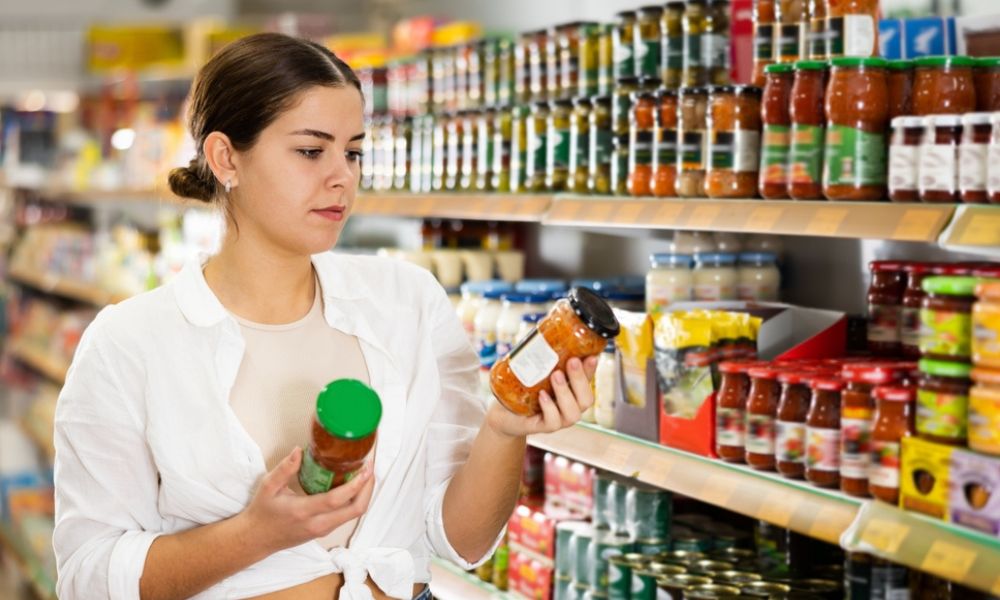 Mulher comparando dois potes de molho de tomate em um corredor de supermercado.