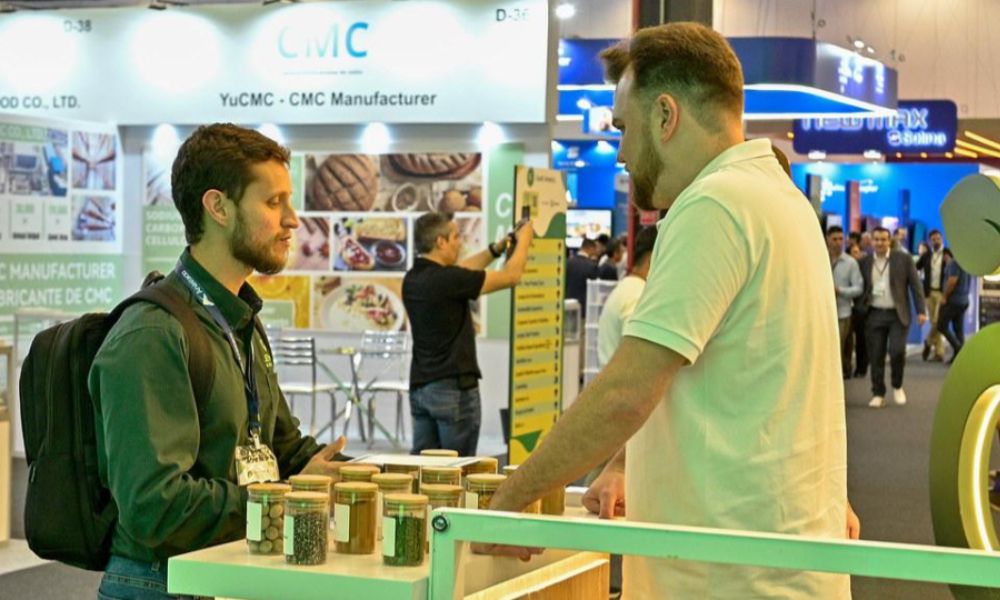 Dois homens conversando em um estande de feira de negócios, com potes de amostras de produtos exibidos sobre o balcão.