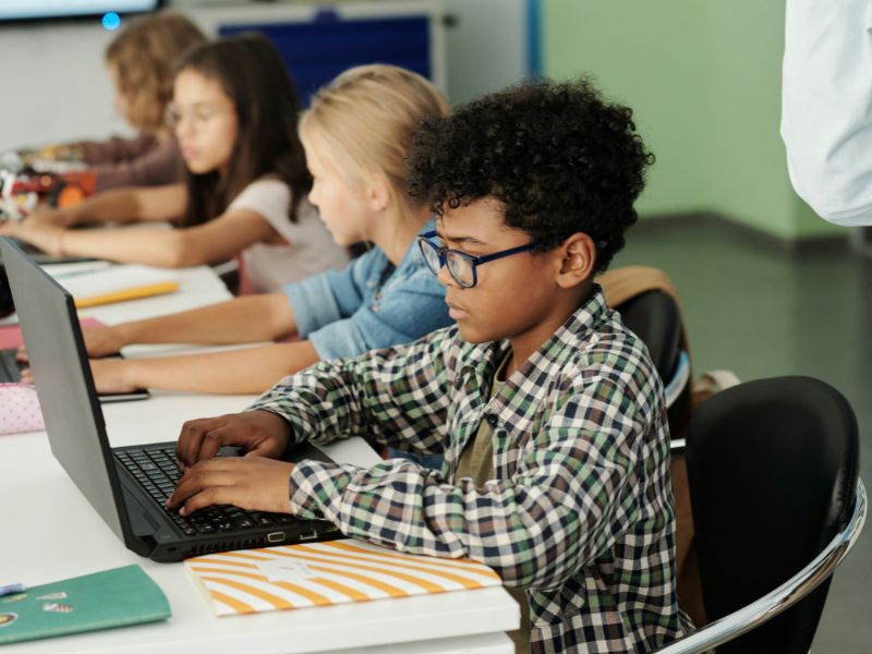 A imagem mostra um grupo de crianças em uma sala de aula moderna, concentradas em atividades com computadores portáteis.