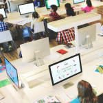 A imagem mostra uma sala de aula moderna, equipada com diversos computadores em mesas amplas e bem iluminadas. Crianças estão sentadas diante dos monitores, participando de atividades educacionais digitais