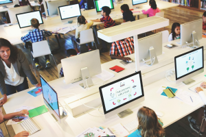 A imagem mostra uma sala de aula moderna, equipada com diversos computadores em mesas amplas e bem iluminadas. Crianças estão sentadas diante dos monitores, participando de atividades educacionais digitais