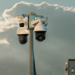 A imagem é uma fotografia de duas câmeras de vigilância tipo dome (cúpula) ou PTZ (Pan-Tilt-Zoom) montadas em um poste, com um fundo de céu parcialmente nublado e um prédio embaçado no canto inferior direito.