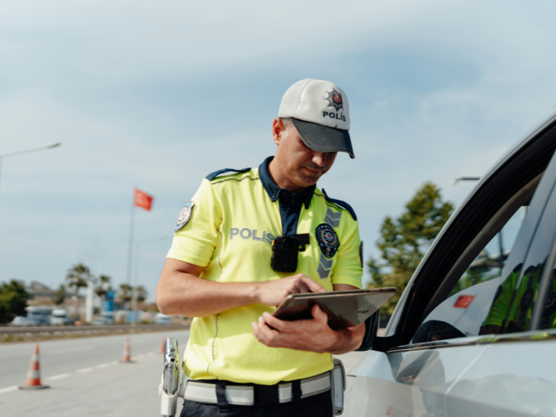A imagem mostra um oficial de polícia (ou segurança pública) em serviço, interagindo com um dispositivo digital próximo a um veículo.