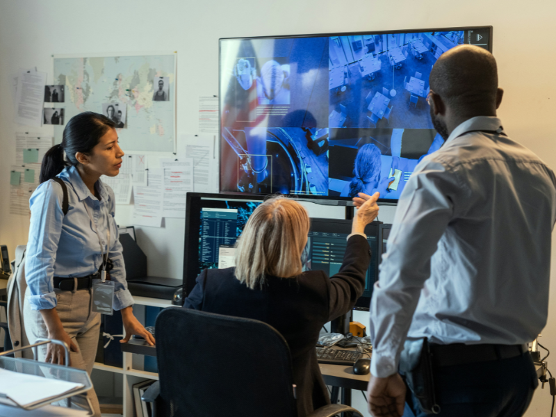 A imagem retrata uma cena em uma sala de monitoramento, investigação ou centro de comando, envolvendo três profissionais que analisam informações em telas de computador e monitores.
