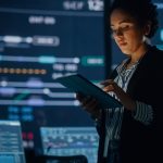 Uma jovem funcionária pública multirracial utiliza um tablet no centro de monitoramento e controle de sistemas. Ao fundo, seus colegas de trabalho estão em seus postos de trabalho, com diversos monitores exibindo dados técnicos.