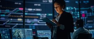 Uma jovem funcionária pública multirracial utiliza um tablet no centro de monitoramento e controle de sistemas. Ao fundo, seus colegas de trabalho estão em seus postos de trabalho, com diversos monitores exibindo dados técnicos.