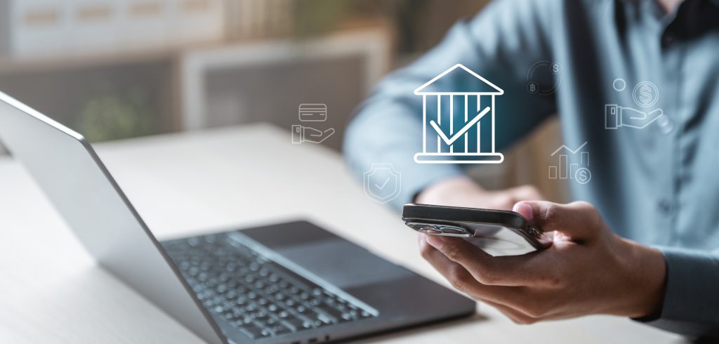 Uma pessoa está usando um laptop e um telefone celular para olhar para uma conta bancária. O notebook está aberto para uma tela com um logotipo do banco