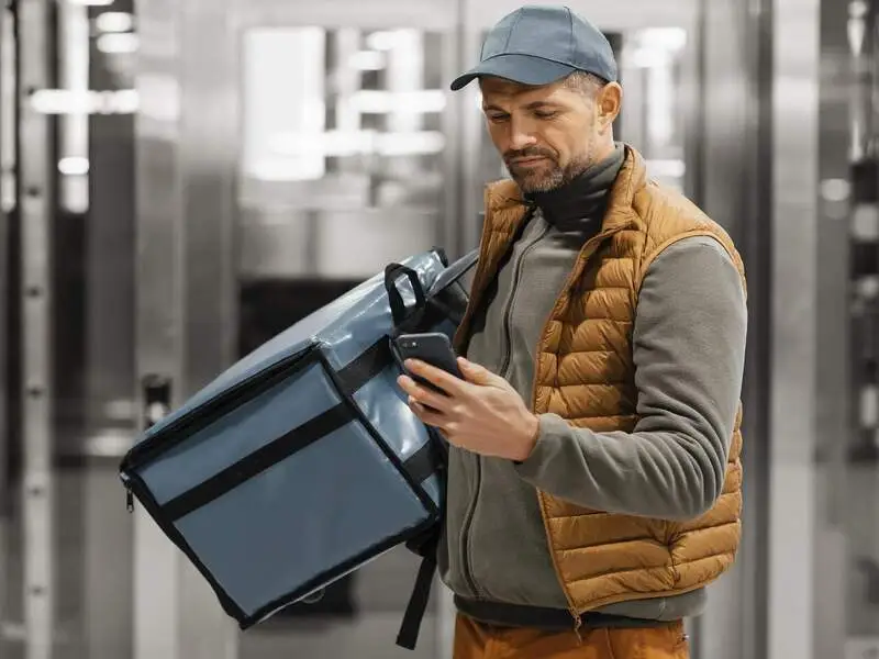 funcionário de boné, bolsa térmica e carregando um celular fazendo a entrega de produto perecível