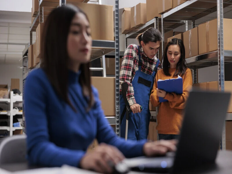 funcionária de camisa branca analisa como funciona um notebook, enquanto outros dois funcionários visualizam uma prancheta