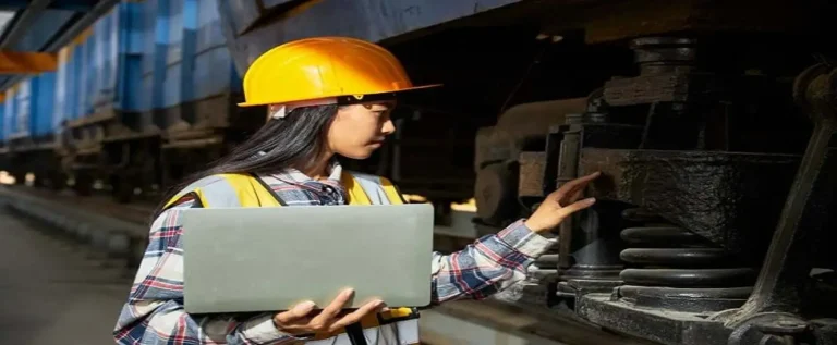 funcionária de capacete amarelo e notebook faz a manutenção ferroviária ao checar os trilhos