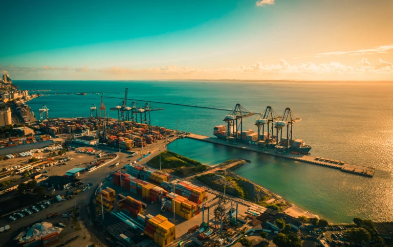 Vista aérea de um porto com contêineres e guindastes, destacando a atividade comercial e o movimento do transporte marítimo ao pôr do sol.