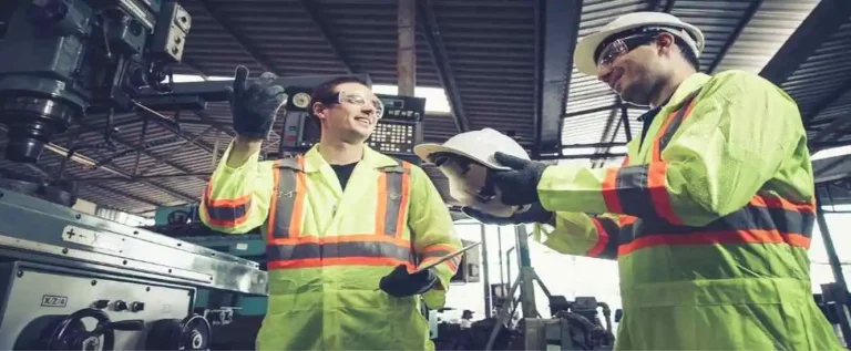 dois funcionários de casaco amarelo e capacete discutindo sobre os fatores humanos na prevenção de riscos