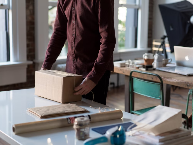 A imagem mostra uma pessoa em pé, embalando uma caixa de papelão sobre uma mesa branca em um ambiente de trabalho iluminado por luz natural.