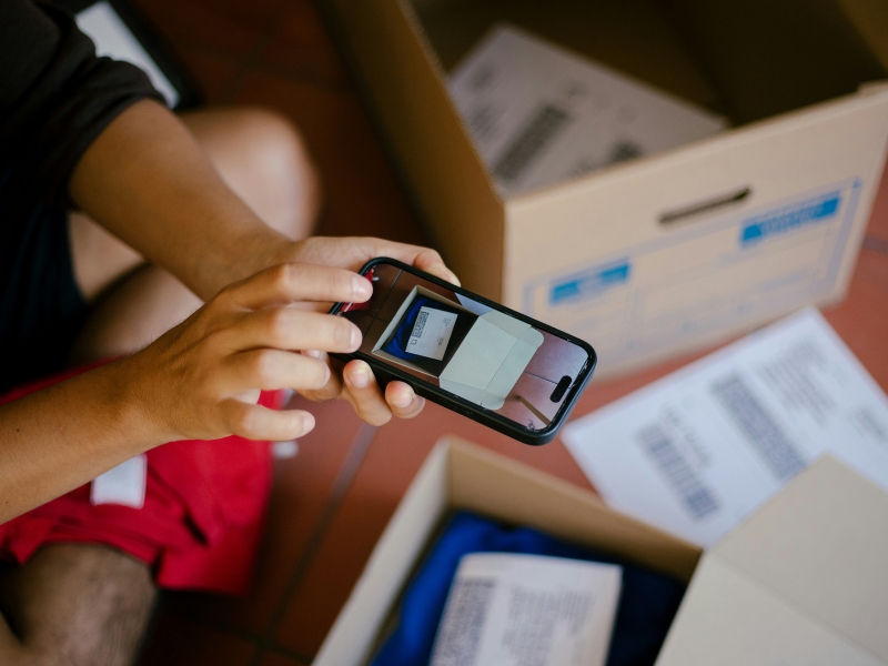 A imagem mostra uma pessoa usando um smartphone para fotografar ou escanear um documento com código de barras, provavelmente relacionado a envios ou logística. Ao redor, há caixas de papelão abertas contendo papéis e itens embalados, sugerindo um processo de organização, despacho ou controle de estoque.