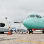 Avião de cor verde claro estacionado no aeroporto, com veículo de carregamento de combustível ao lado, em um dia nublado.