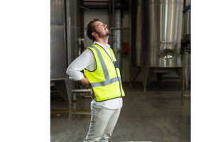A imagem mostra um homem em um ambiente industrial, usando um colete de segurança amarelo com faixas refletivas e uma camisa branca. Ele está de pé, com uma das mãos apoiada na região lombar e a cabeça inclinada para trás, aparentando sentir dor nas costas.