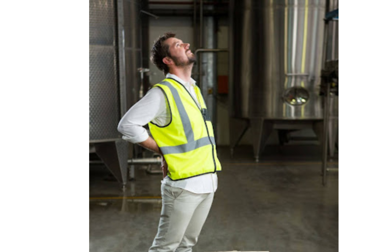 A imagem mostra um homem em um ambiente industrial, usando um colete de segurança amarelo com faixas refletivas e uma camisa branca. Ele está de pé, com uma das mãos apoiada na região lombar e a cabeça inclinada para trás, aparentando sentir dor nas costas.