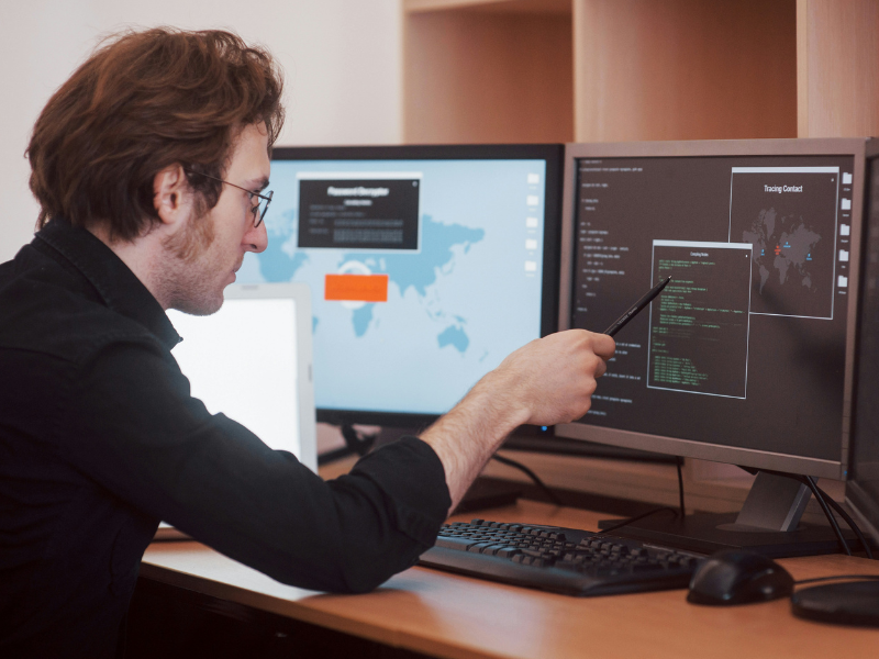 A imagem é uma fotografia que mostra um homem trabalhando intensamente em uma estação de trabalho com múltiplos monitores, sugerindo atividades de programação, análise de dados ou segurança cibernética.