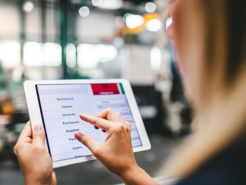 A imagem é uma fotografia em close-up que foca nas mãos de uma pessoa (provavelmente uma mulher, devido à parte do cabelo visível) interagindo com um tablet digital, sugerindo a utilização de um software de gestão em um ambiente de logística ou industrial.
