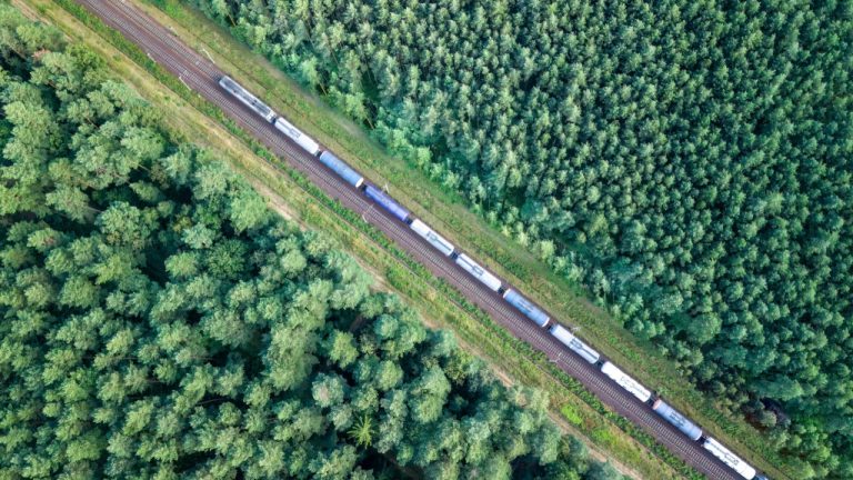 Trem de carga transportando petróleo através da floresta de coníferas virgem ao pôr do sol, vista aérea de cima para baixo.