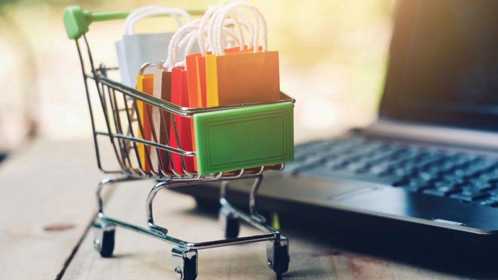 Carrinho de compras de brinquedo em close-up com uma pequena sacola e um computador sobre a mesa, tecnologia de comércio eletrônico e negócios online, pandemia global e crise econômica, novo normal, conceito de ficar e trabalhar em casa