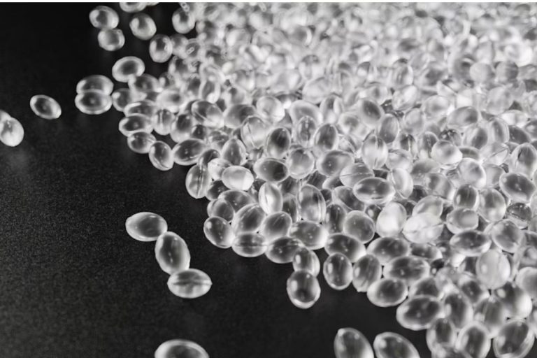 A imagem mostra uma grande quantidade de pellets de resina plástica transparente, que são pequenas esferas ou grânulos utilizados como matéria-prima na indústria de transformação de plásticos.