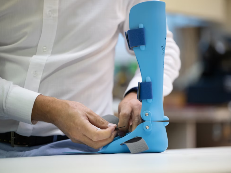 A imagem mostra uma pessoa ajustando uma órtese ou prótese azul, possivelmente feita por impressão 3D. A órtese é presa com fitas de velcro e parafusos metálicos.