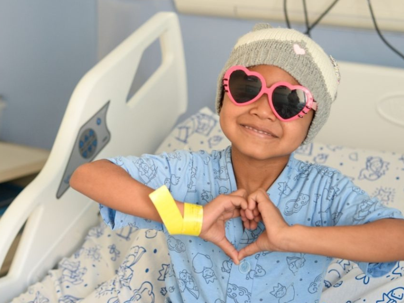 Criança feliz no hospital fazendo um coração com as mãos, usando óculos de sol de coração rosa e chapéu de lã.