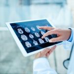Médico analisando exames de ressonância cerebral em tablet em consultório, representando tecnologia médica avançada e diagnóstico por imagens. A imagem reflete sobre gêmeos digitais na saúde.