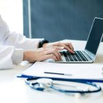 Imagem de um profissional de saúde digitando em um laptop, com uma prancheta e estetoscópio na bancada, simbolizando o ecossistema de saúde no Brasil.